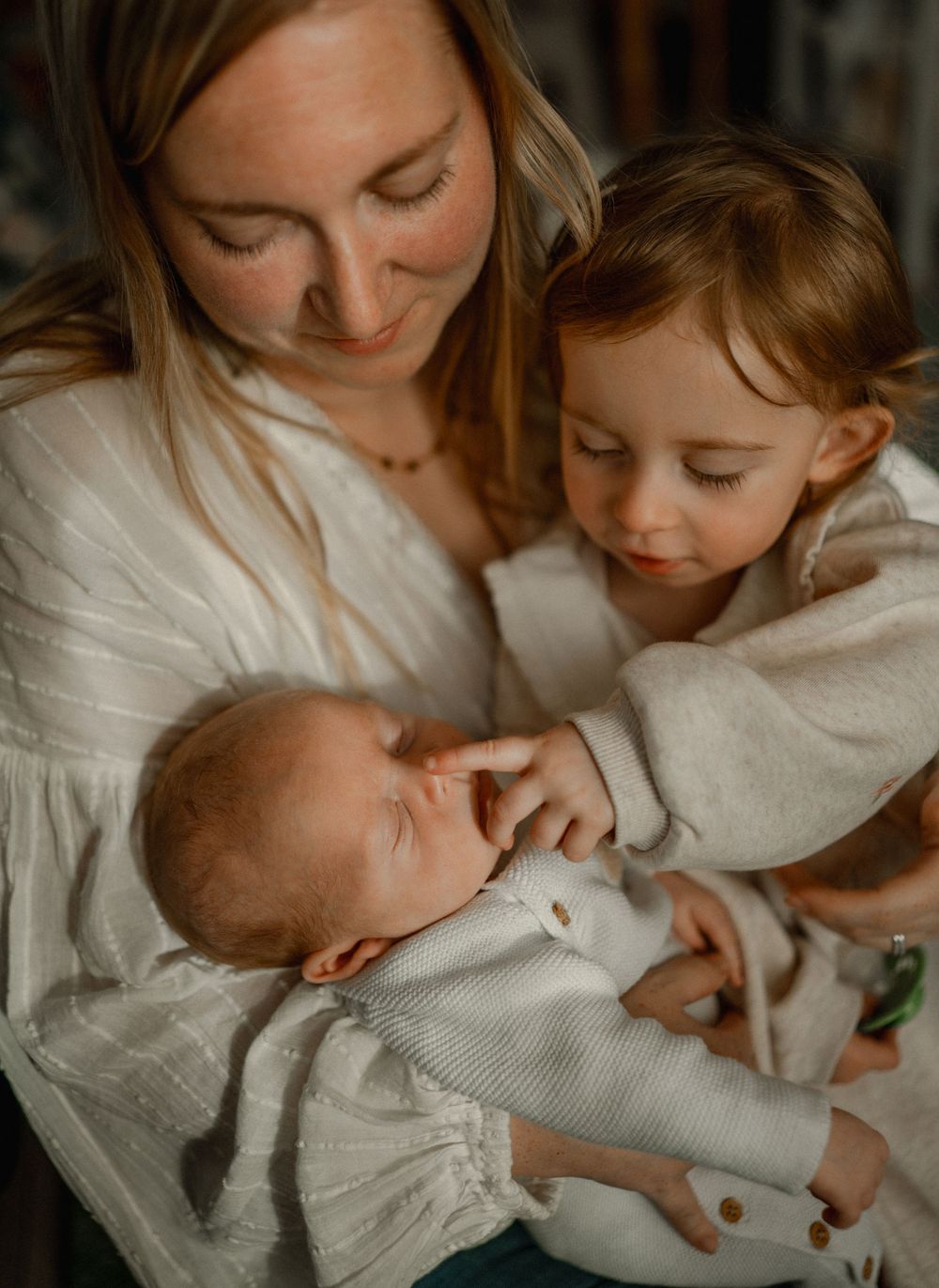 A mother tenderly holds her newborn baby while her toddler gently touches the infant, all dressed in soft white clothing.