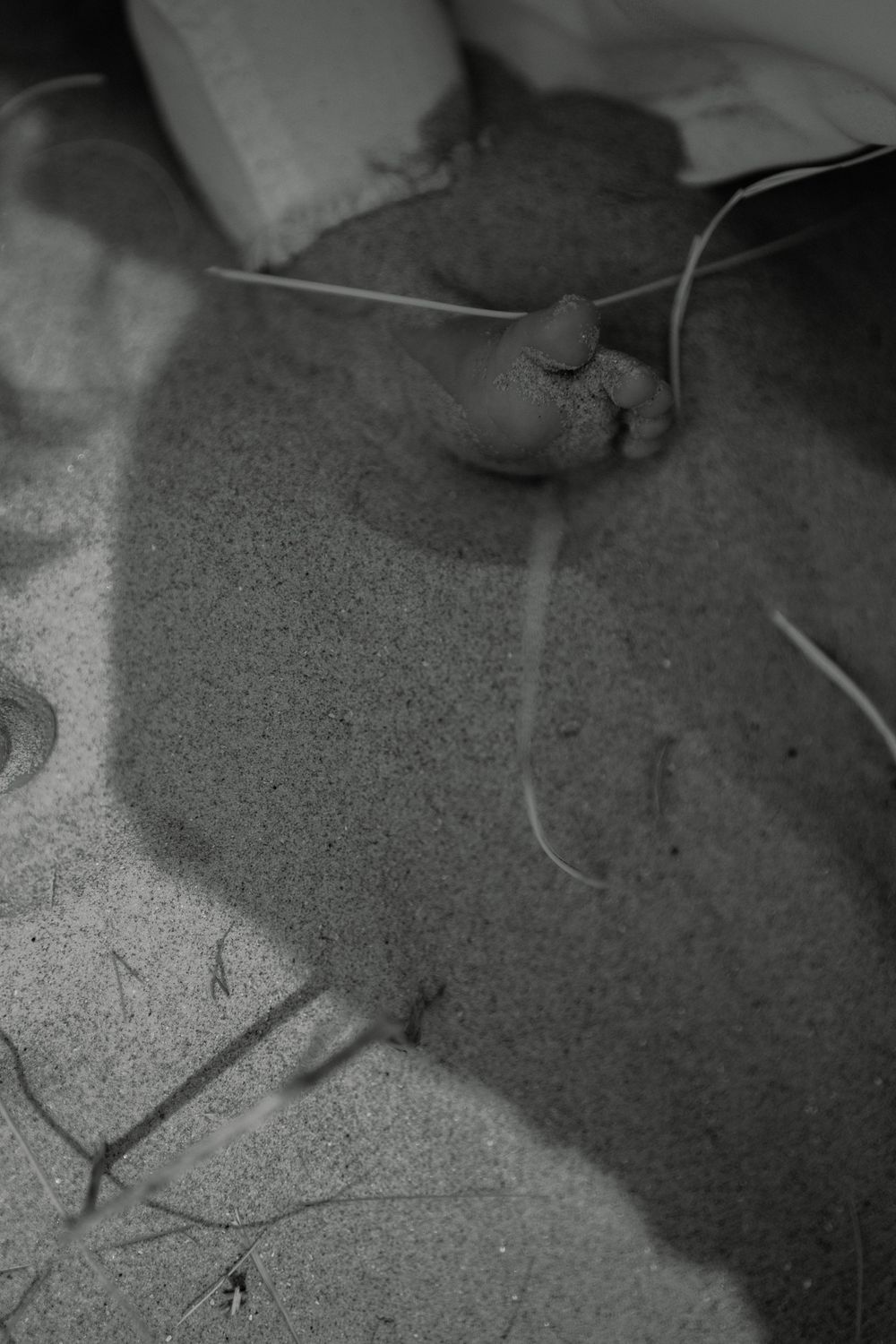 Black and white close-up of bare baby feet resting on sandy beach ground with scattered grass blades.