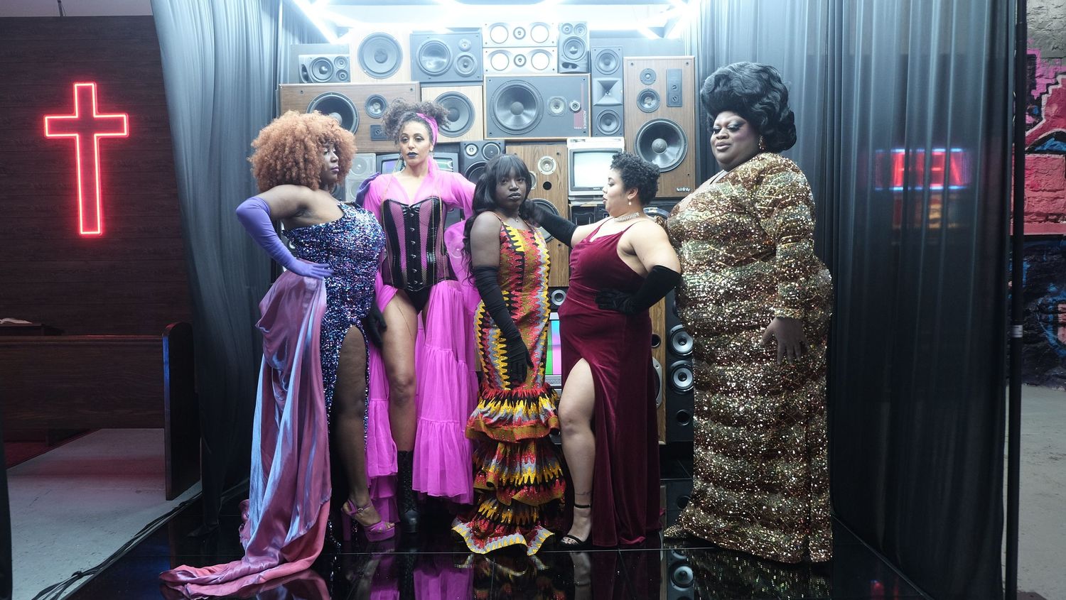 Colorful backstage scene with elaborate costumes displayed against black backdrop with neon cross.
