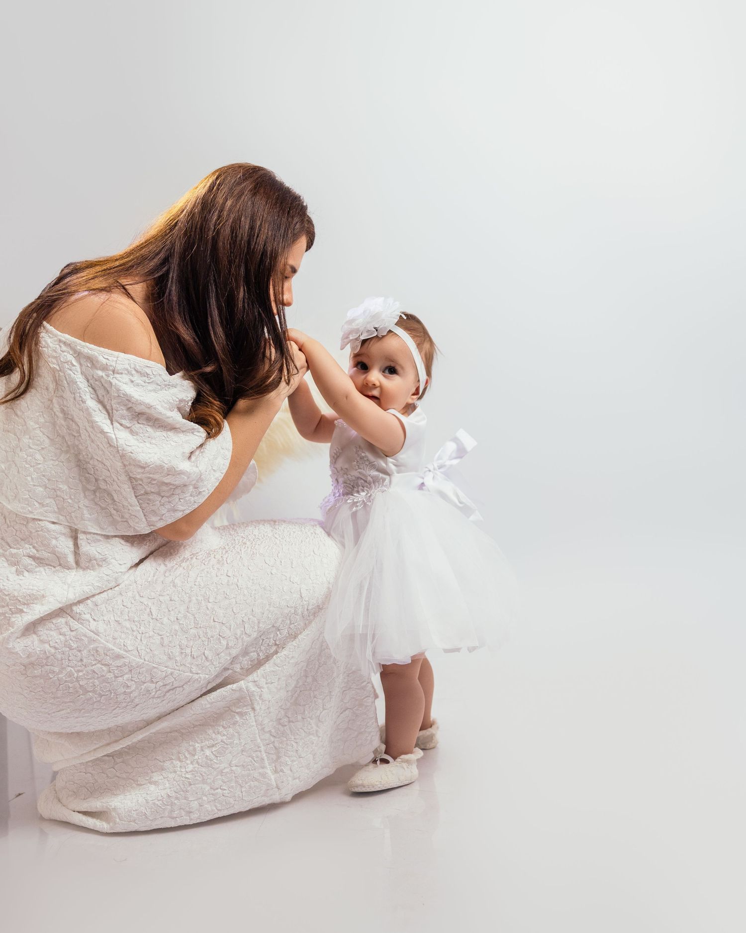 Mother in white dress interacting with baby girl in white tutu dress in studio.