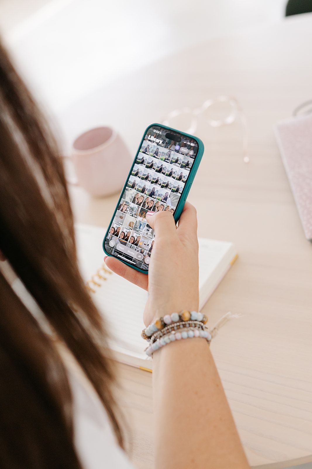 Hands holding a smartphone displaying a photo grid interface with turquoise blue nail polish against a white desk surface.
