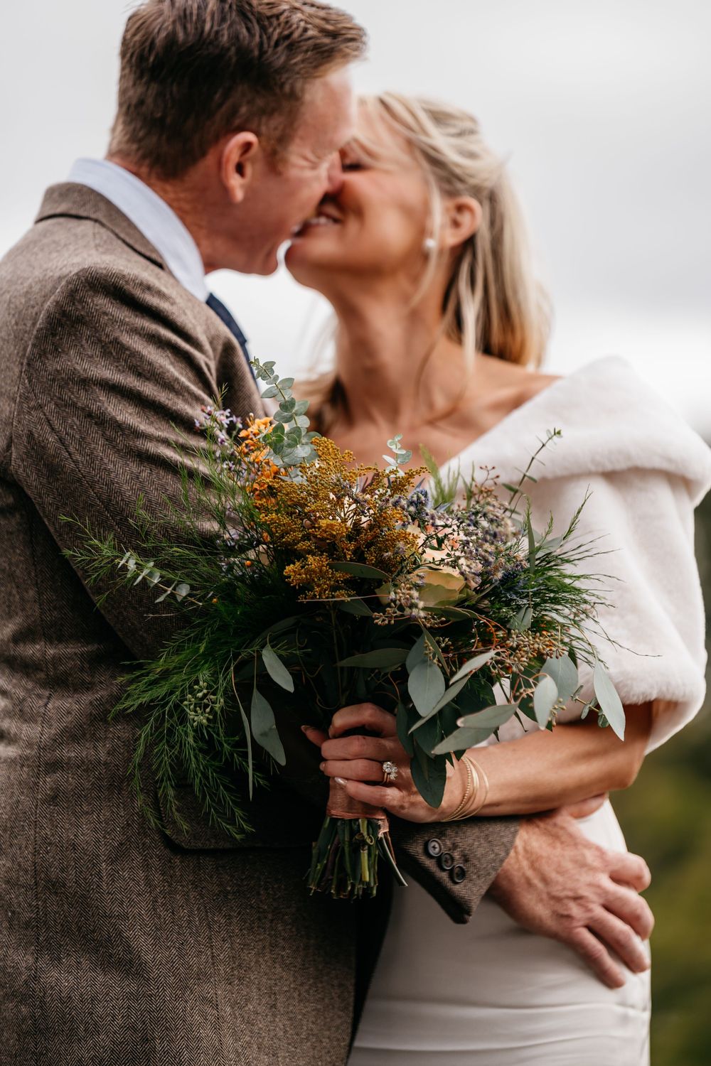 A romantic wedding couple embraces while holding a rustic bouquet with greenery and orange flowers against a light background.