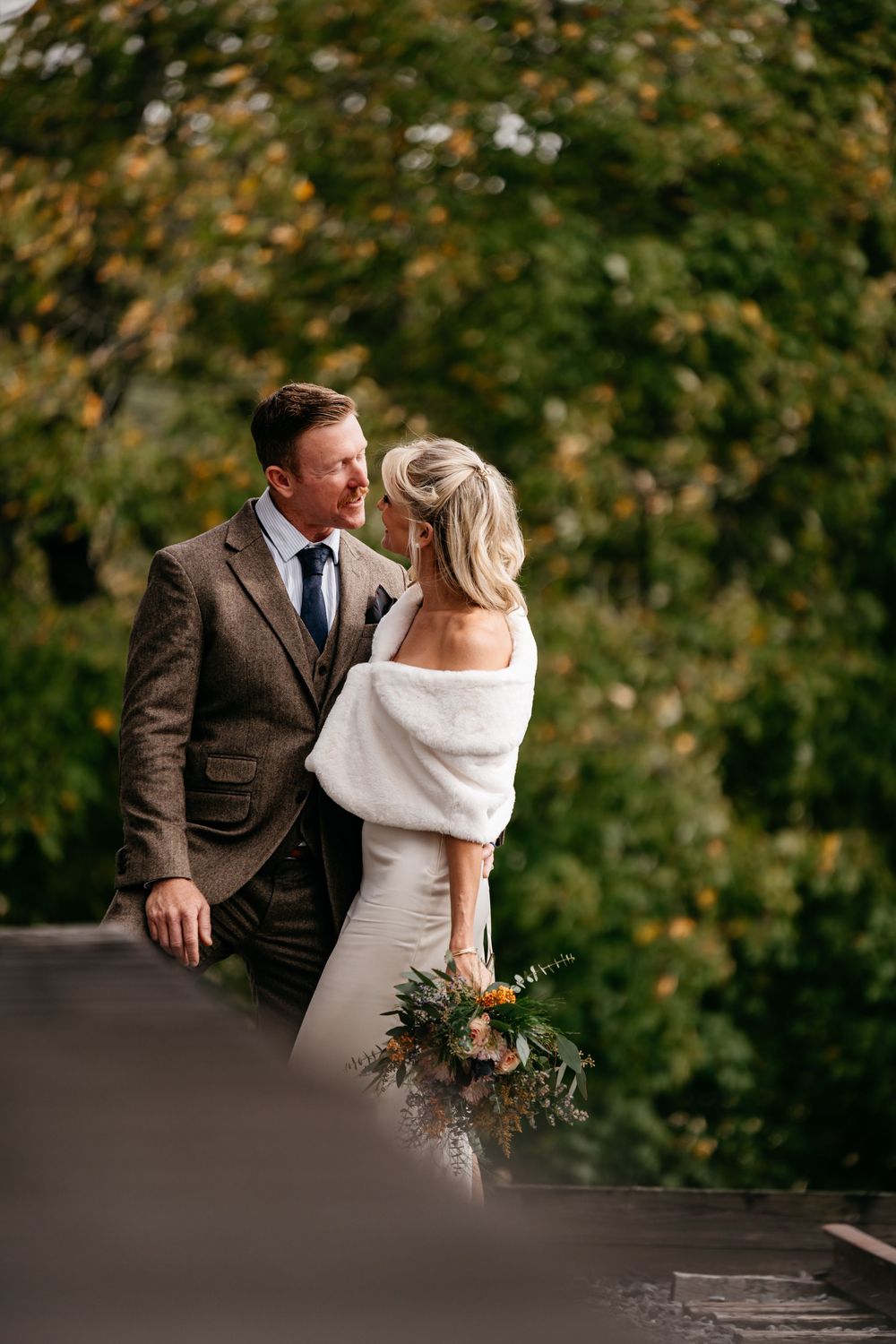 Couple in wedding attire share an intimate moment on a terrace with autumn foliage in the background.