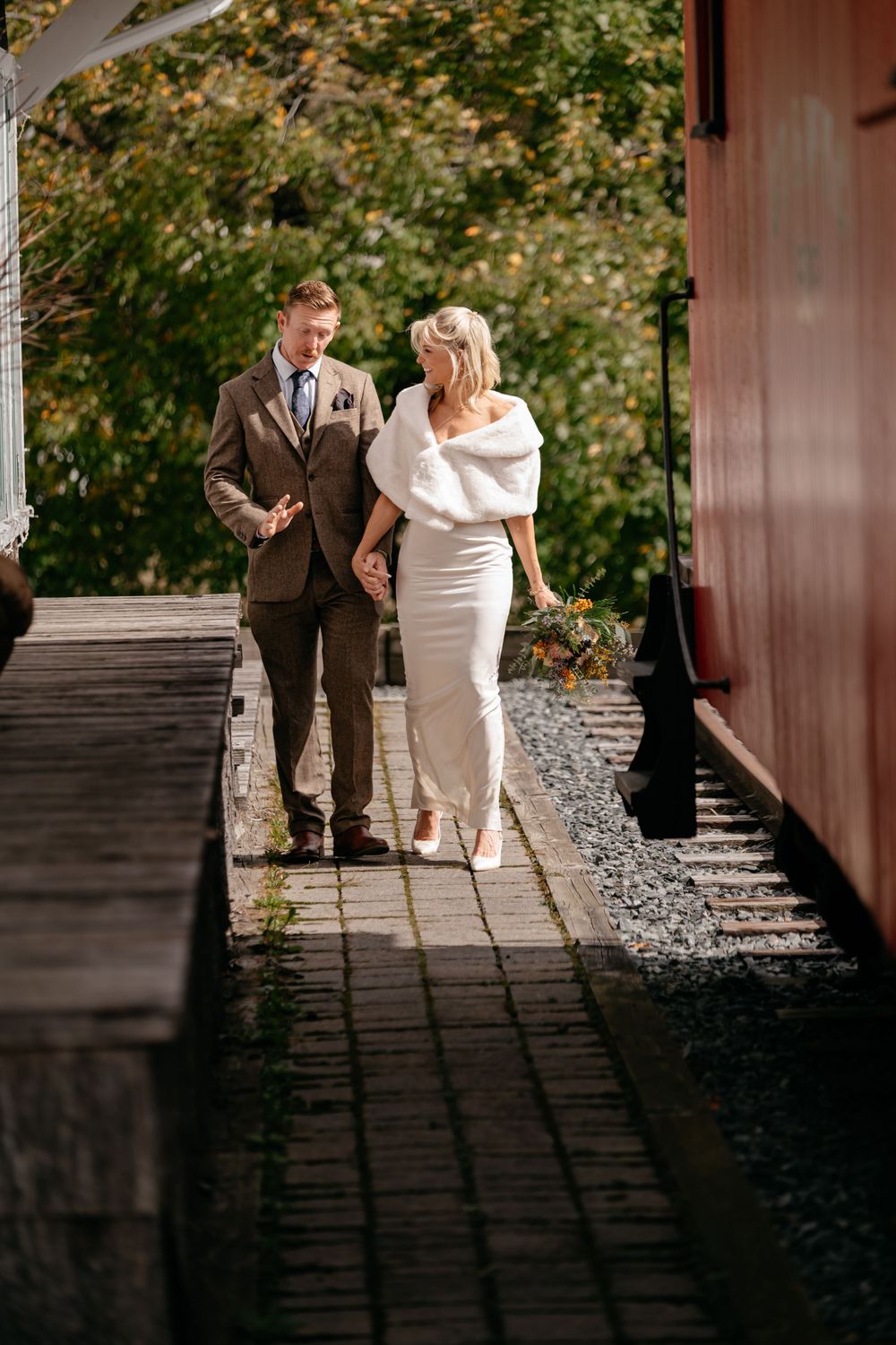 A couple in wedding attire walking together on a stone path beside a rustic wooden building on an autumn day.