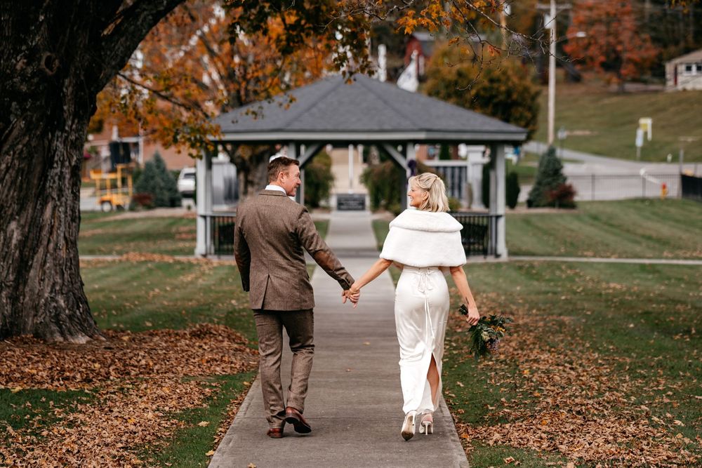 A couple walks together through a park filled with fallen autumn leaves on a sunny day.