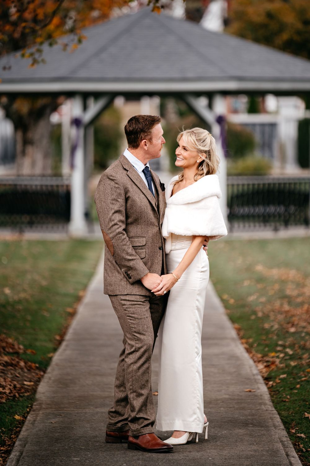 A bride and groom share a romantic moment on a sidewalk lined with autumn leaves during their wedding photoshoot.