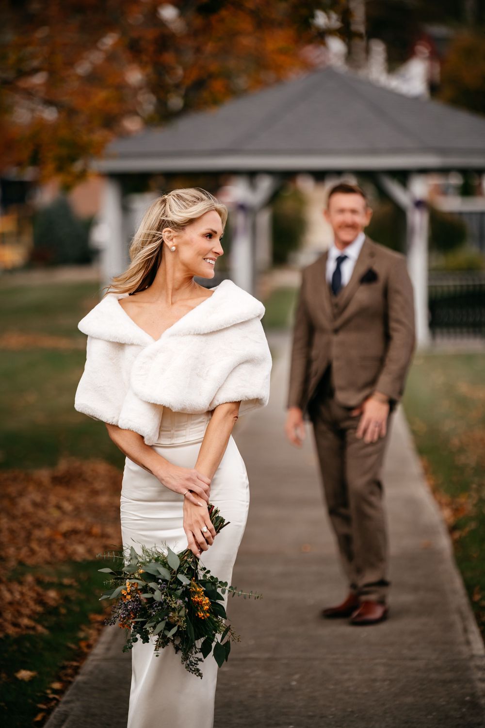 A bride in a white dress holds a bouquet while walking along a path with autumn leaves and trees in the background.