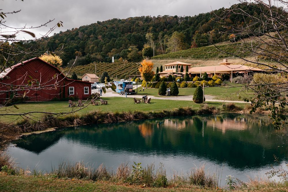 A scenic panoramic view of a lake surrounded by rolling hills, trees, and red barns in a rural countryside setting.