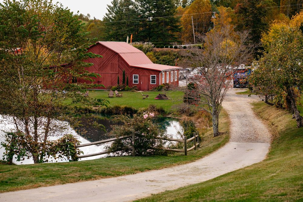 A winding dirt road leads to a red barn nestled among autumn trees beside a tranquil pond in rural New England.