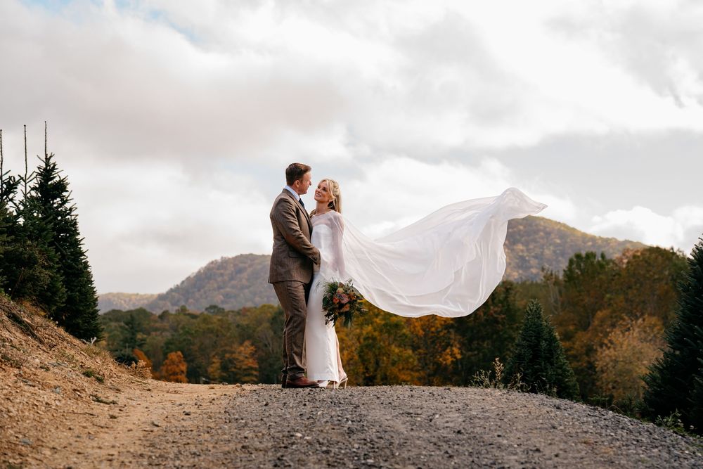 Couple in wedding attire stands on rocky mountain overlook as bride's veil flows dramatically in the wind.