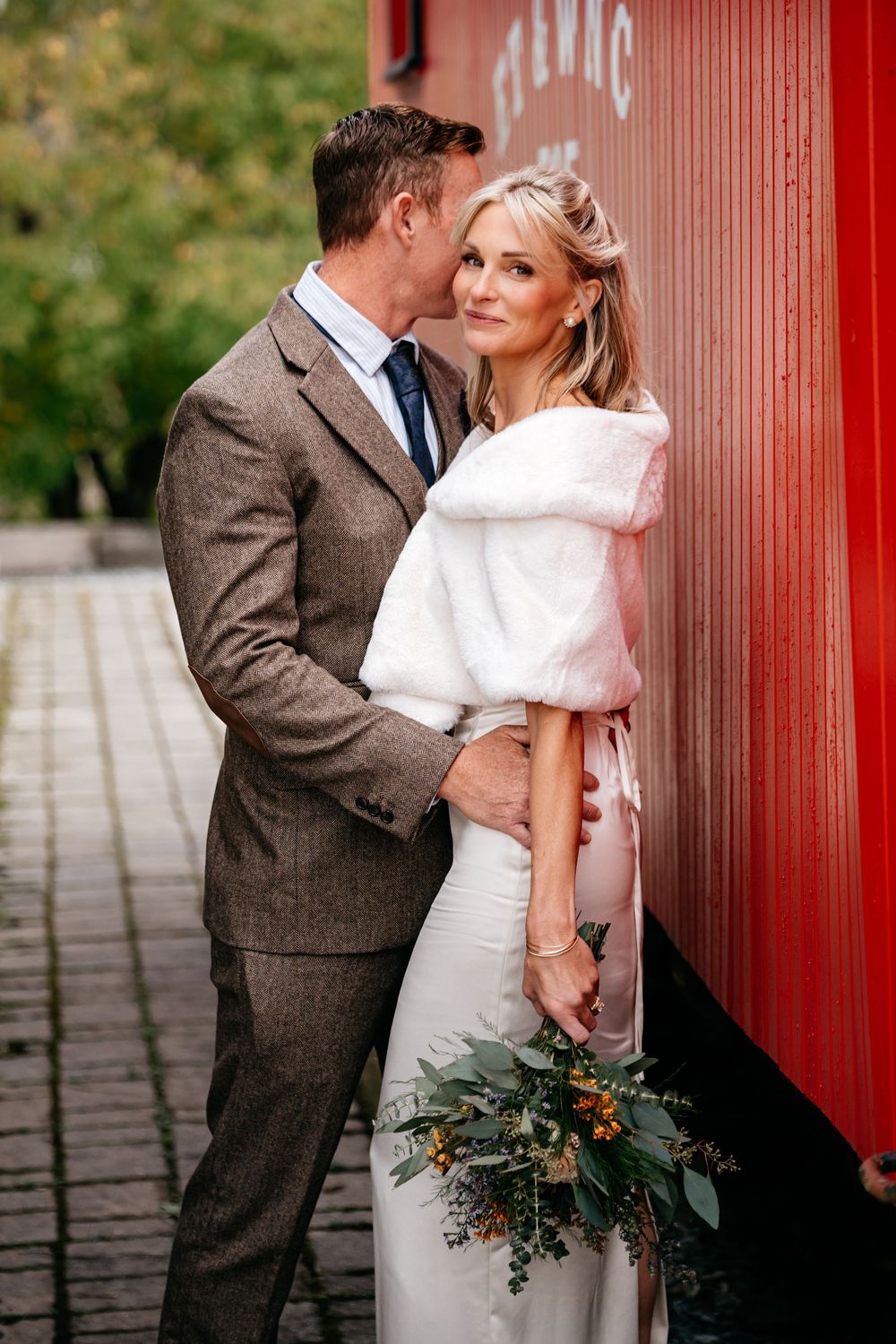 Newlywed couple shares romantic moments against a red wall backdrop with bride holding rustic greenery bouquet.