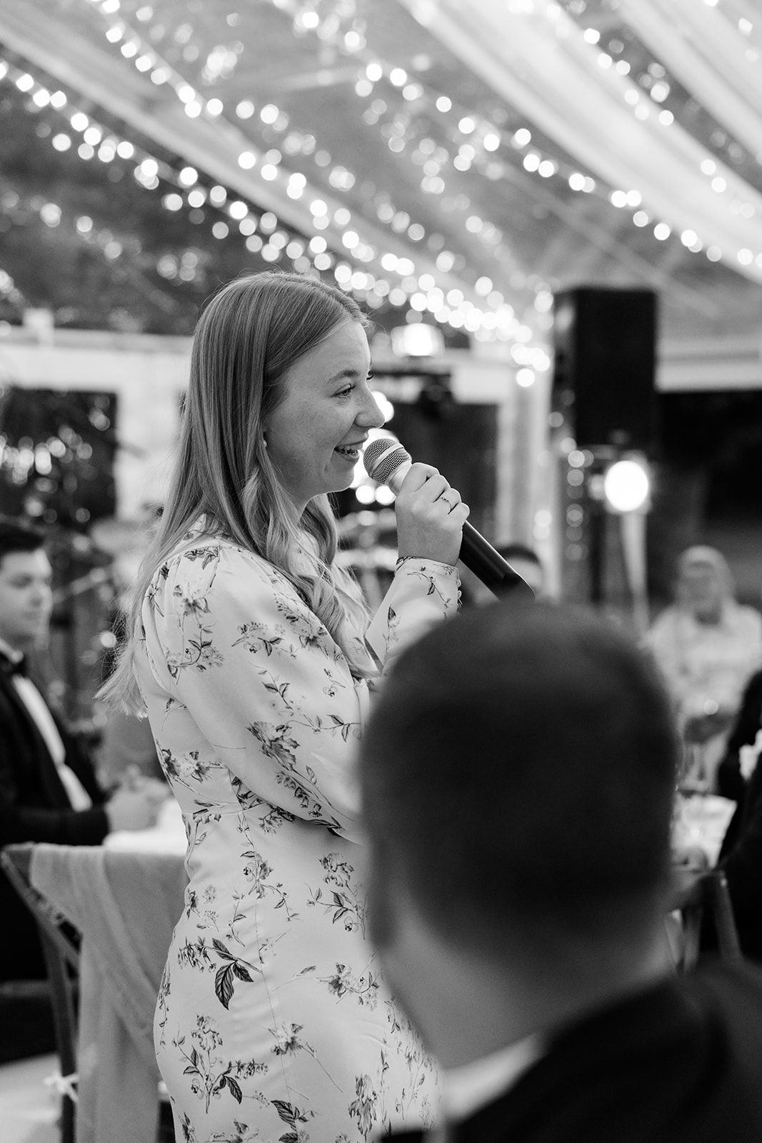 Black and white photo of a person in a floral dress speaking into a microphone at an event with string lights overhead.