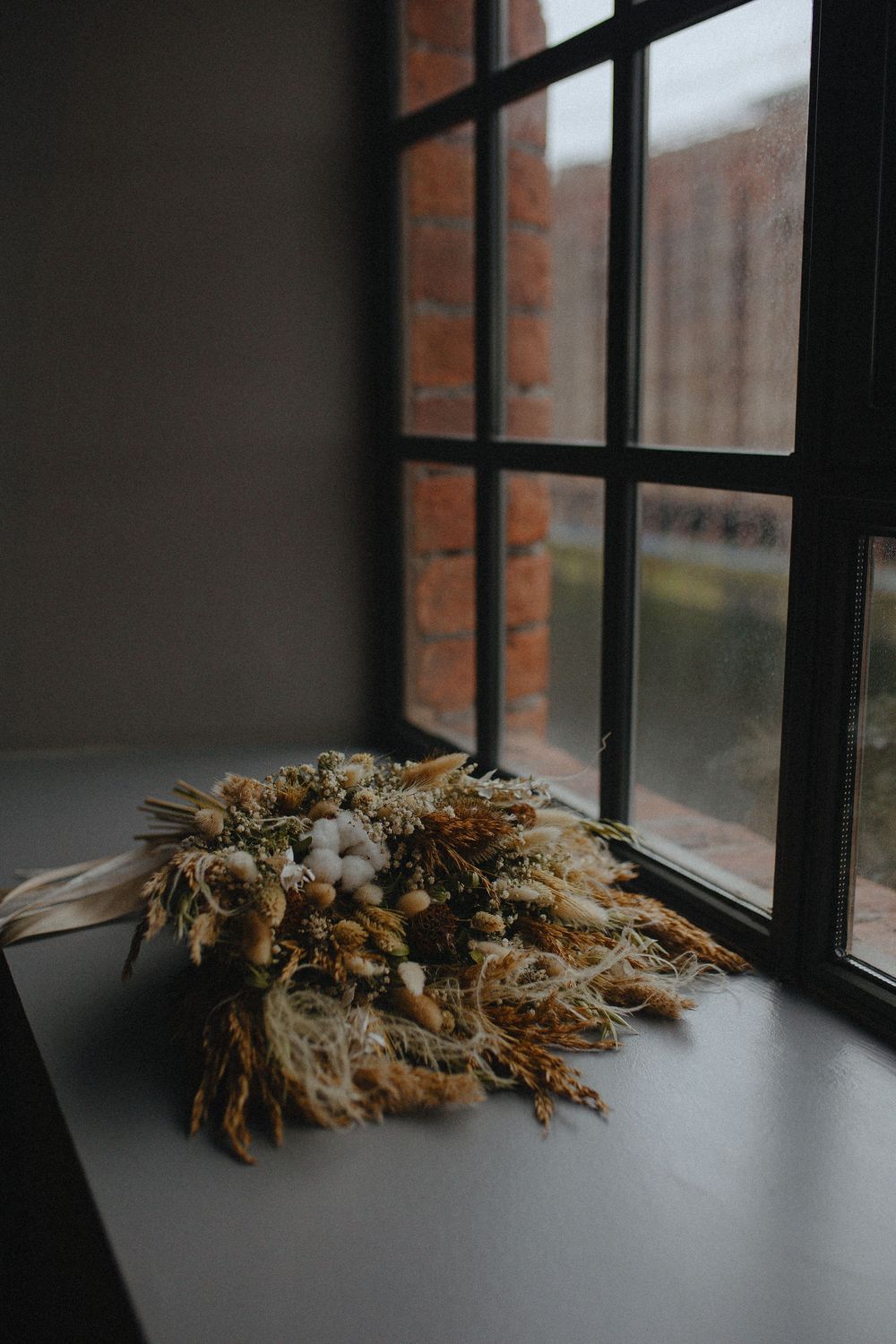 A dried floral arrangement sits on a dark surface near a window with black frames in moody lighting.