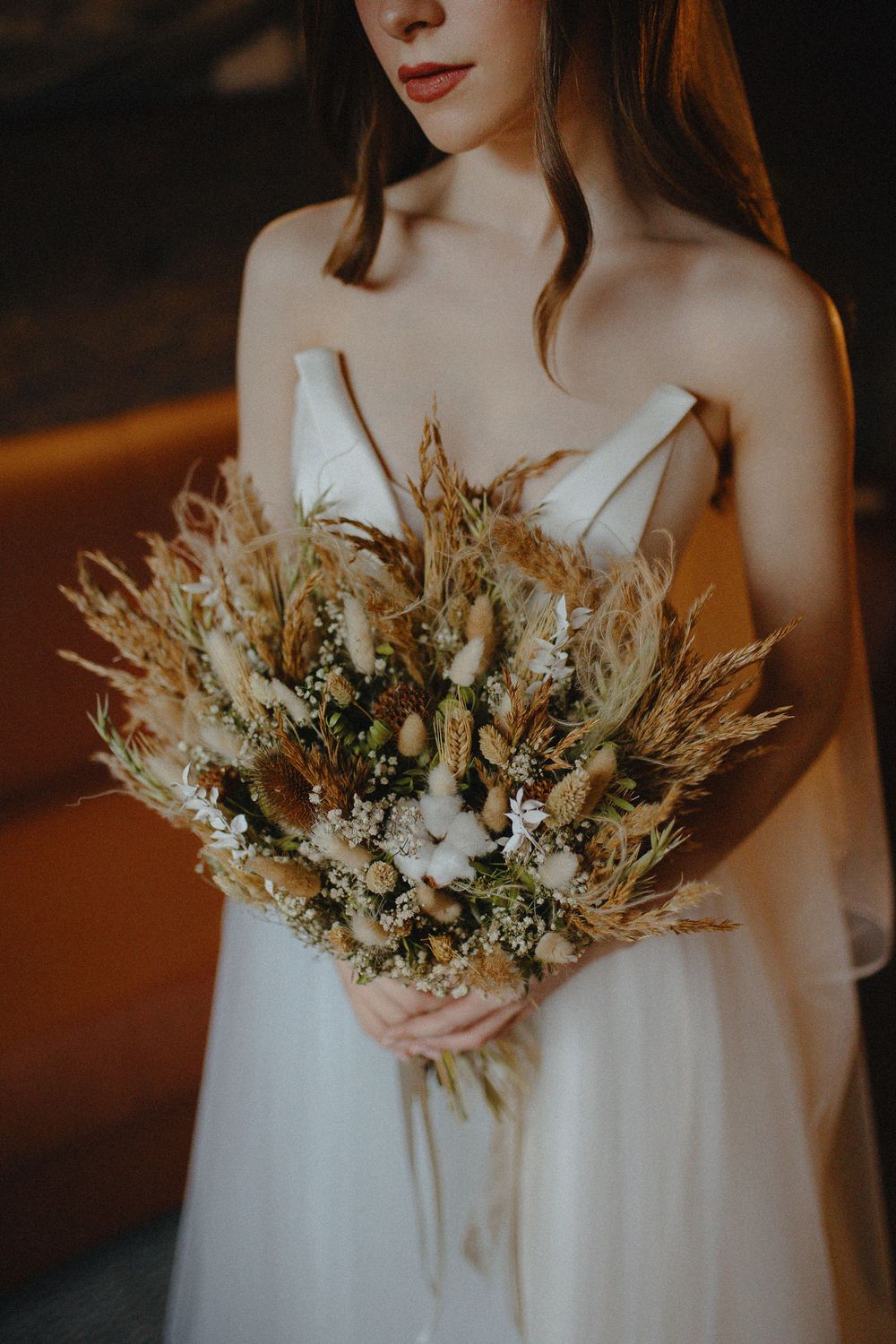 A dried floral wedding bouquet with neutral tones and pampas grass held against a white dress in moody lighting.