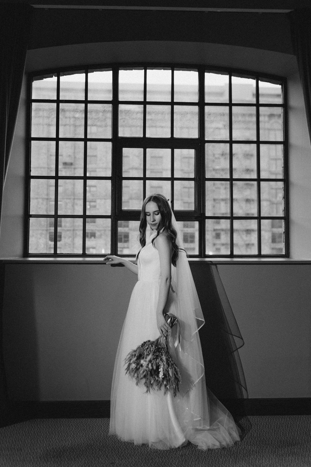 A black and white photograph of a bride in a flowing white gown and veil standing by a large industrial window.