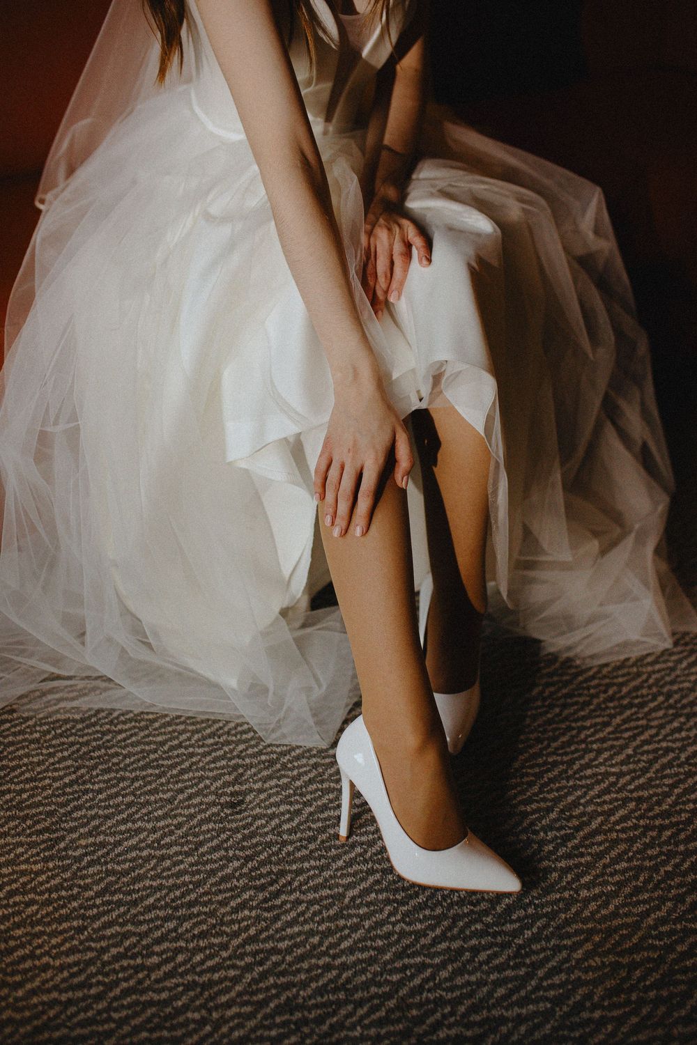 A bride in a white tulle wedding dress puts on elegant white stiletto heels while sitting on gray carpet.