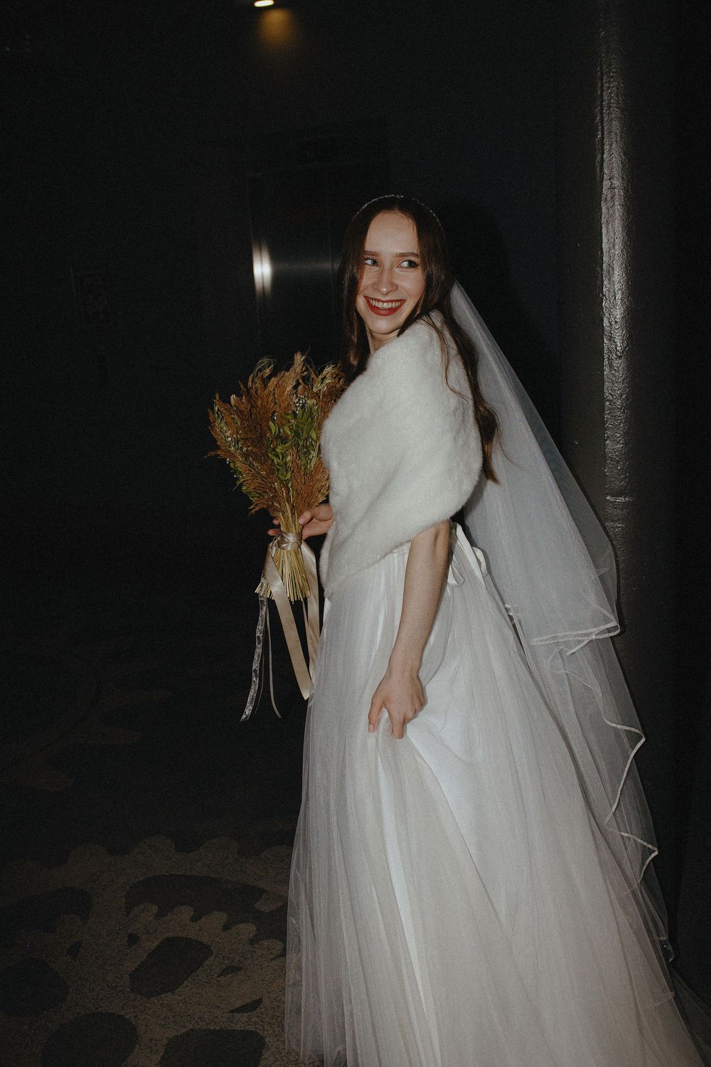 A bride in a white ballgown wedding dress with short sleeves and veil holds a bouquet in a dark setting.