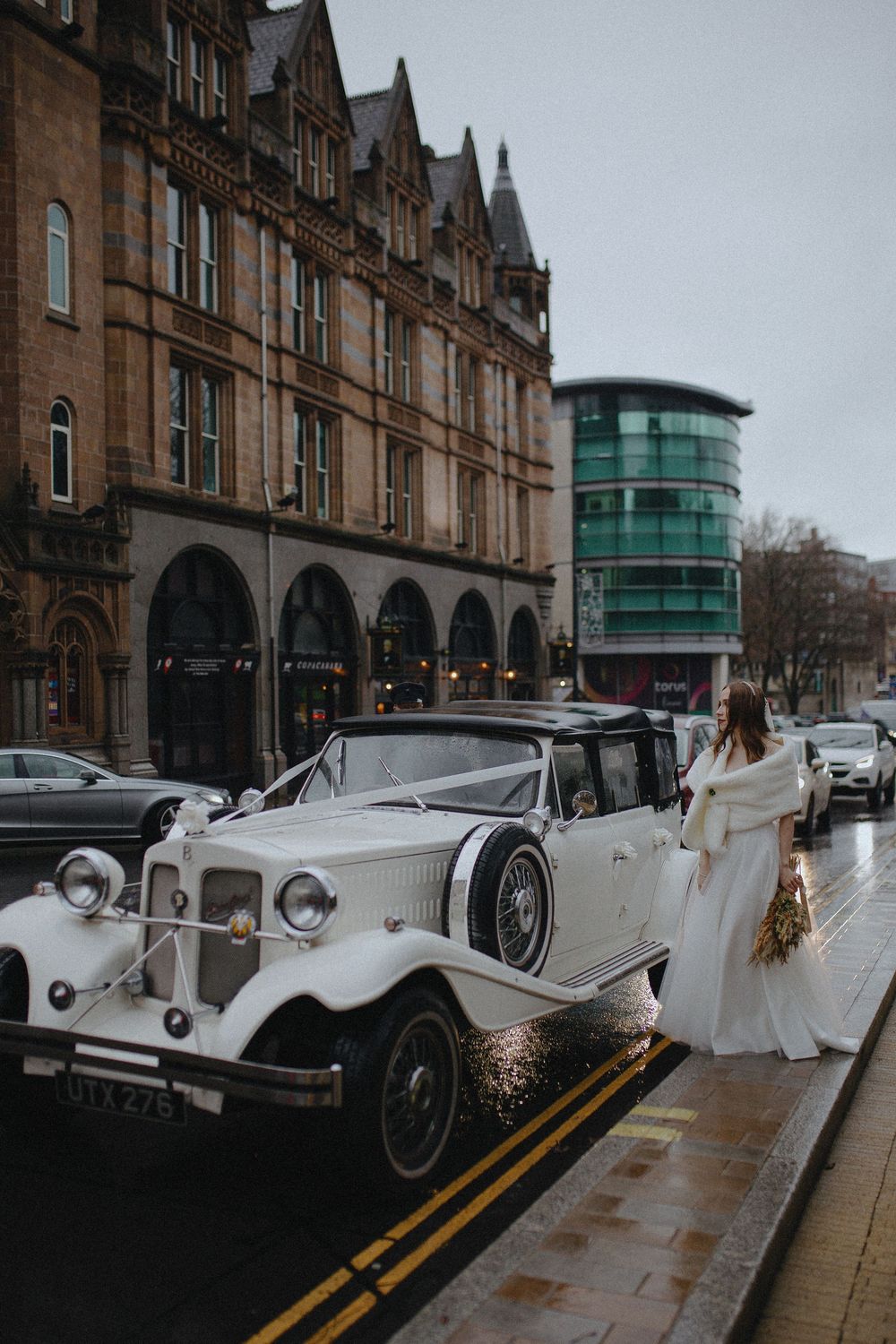 A line of classic white vintage Rolls Royce wedding cars parked along a historic stone building on a rainy day.