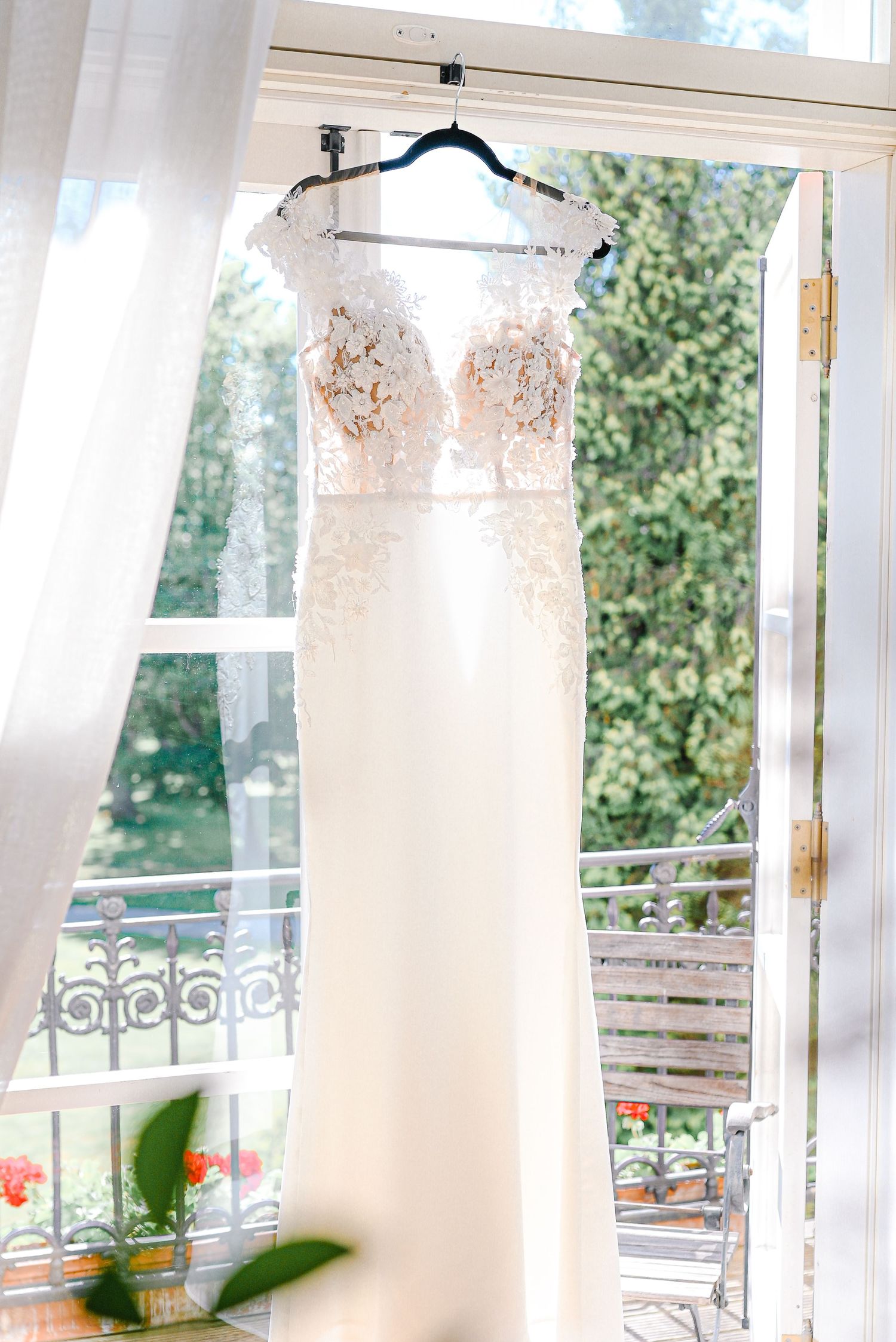 Elegant white lace wedding dress hanging on a hanger in a bright window with garden views.