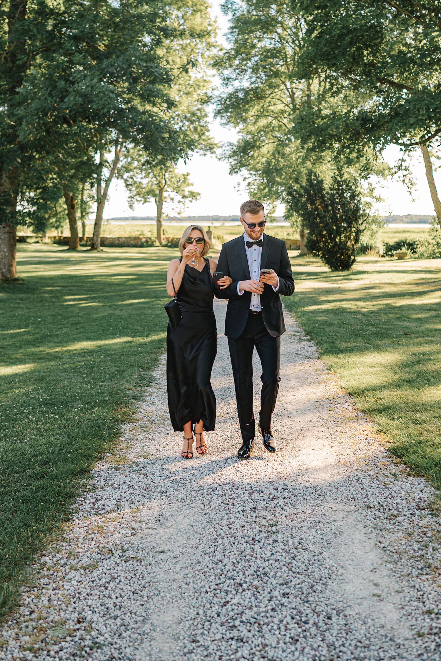 Elegant couple in black formal attire walking on a gravel path surrounded by lush green trees.