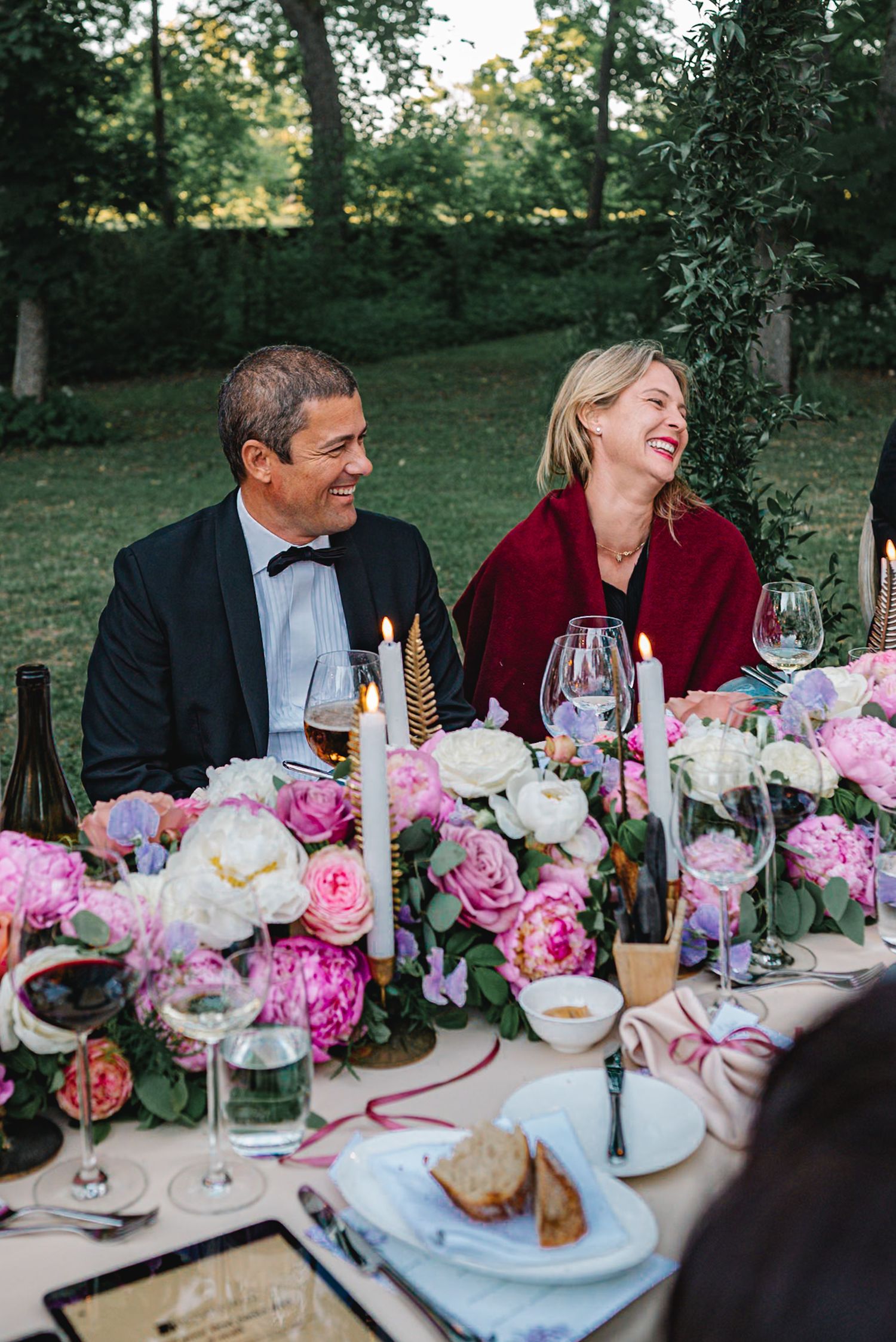 Couple laughing joyfully at an elegant outdoor wedding reception table adorned with pink peonies and roses.