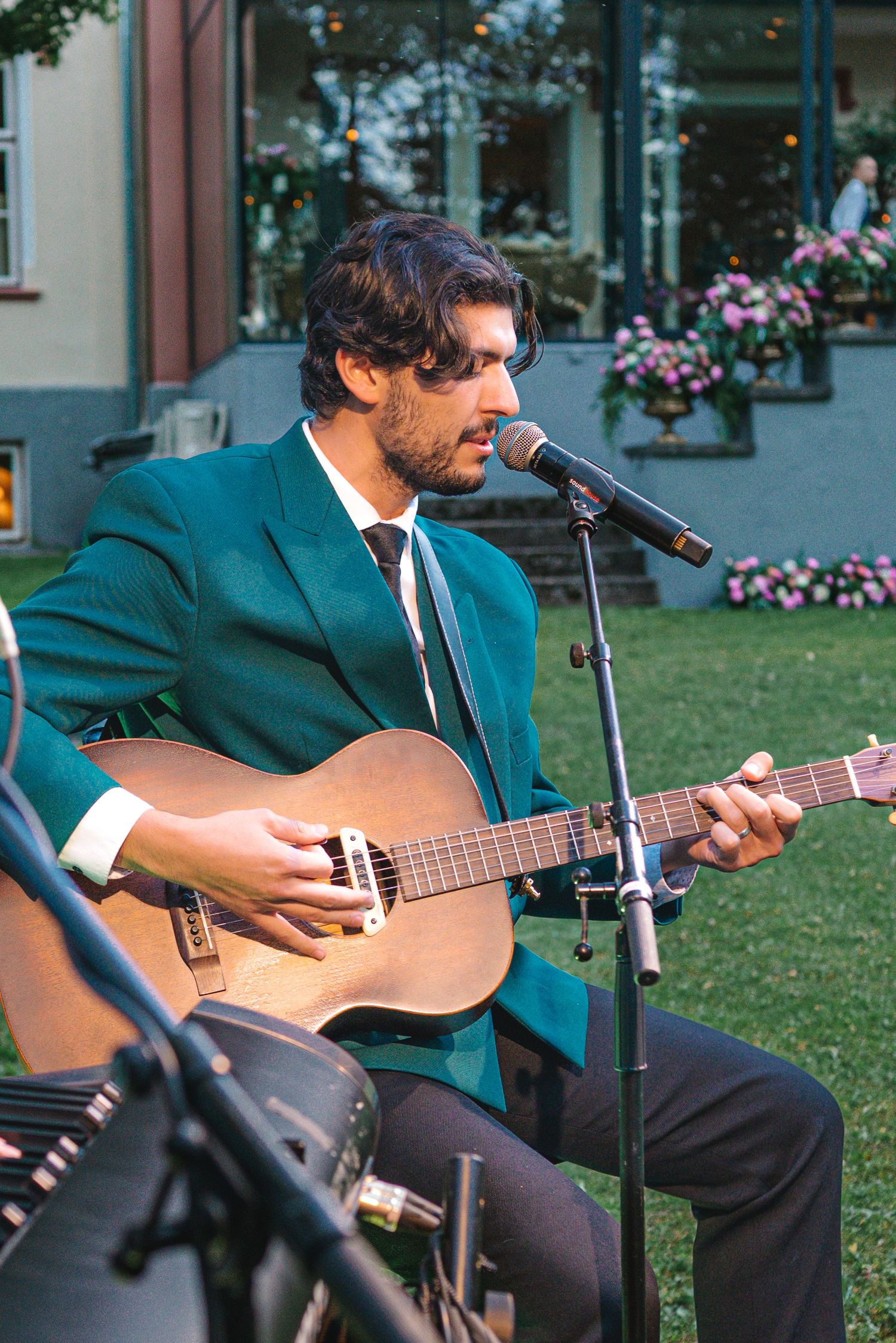 Man in green suit playing acoustic guitar at outdoor microphone stand during garden event.