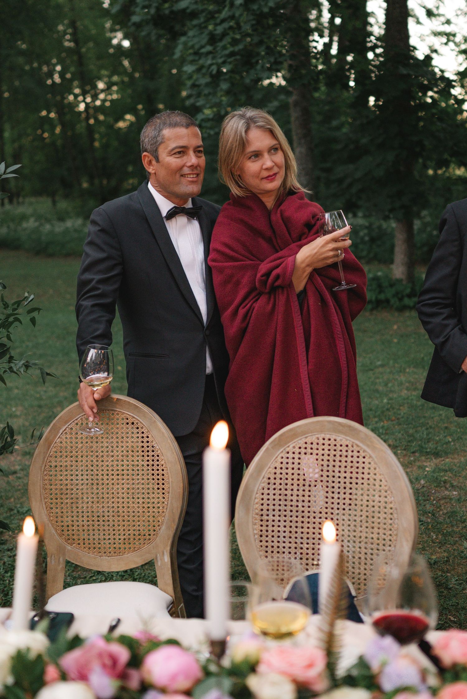 Elegant couple in formal attire toasting with red wine glasses at an outdoor candlelit garden wedding reception.