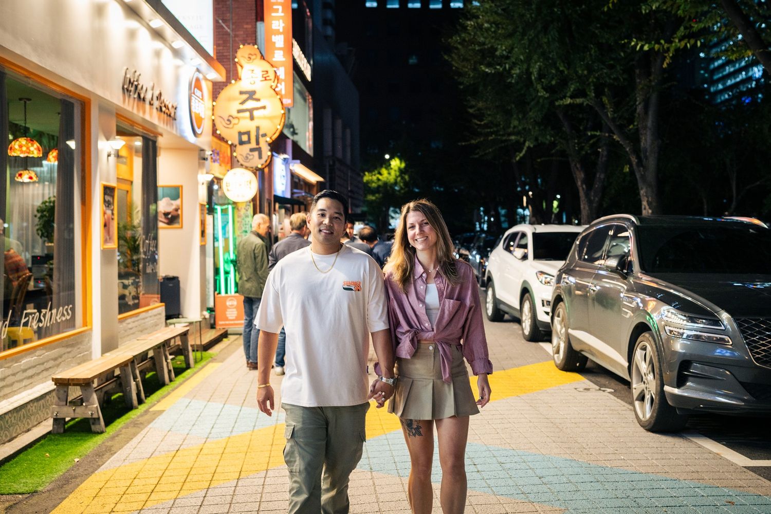 People walk along a brightly lit shopping street at night with storefronts and cars visible.