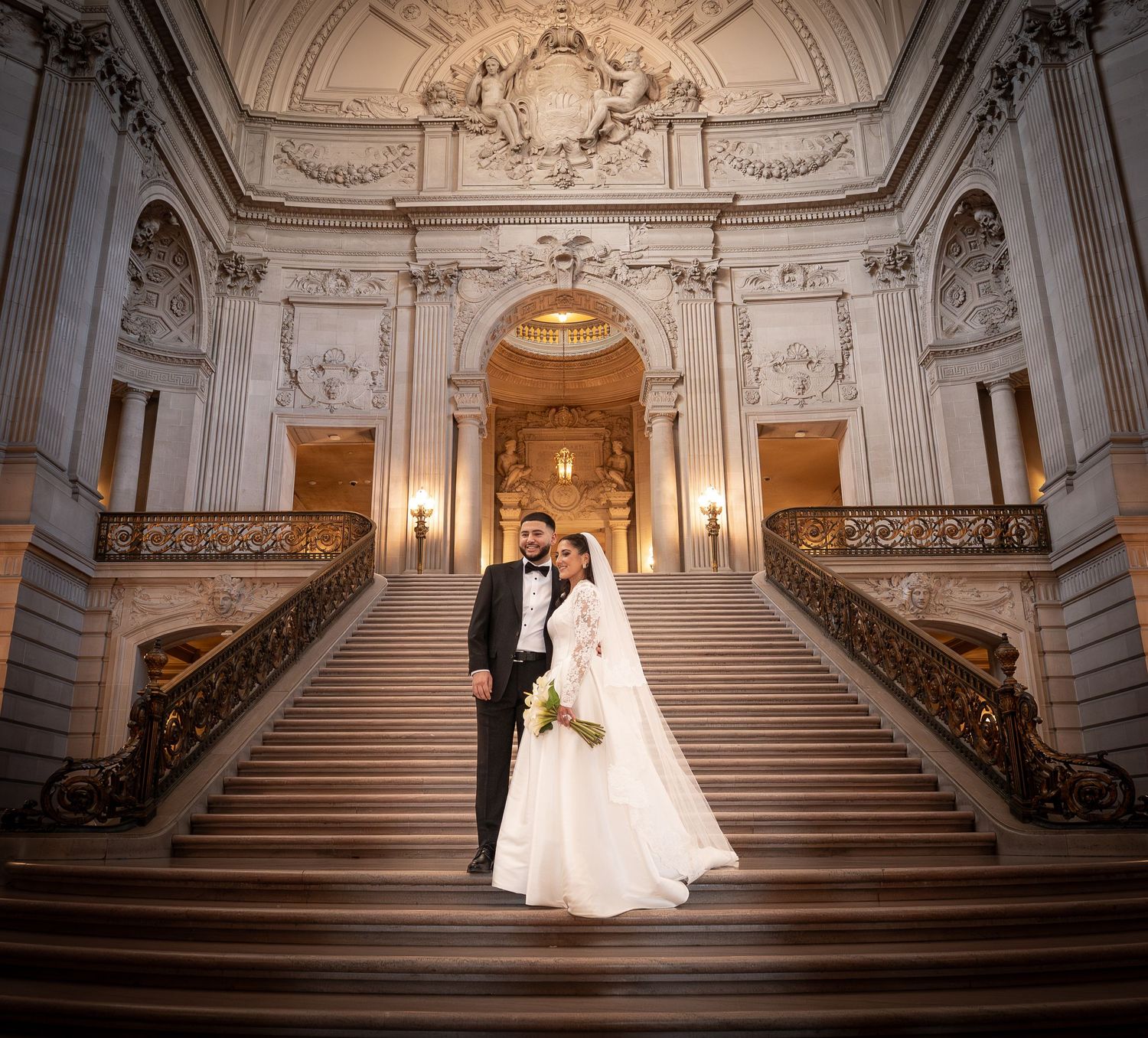A couple in wedding attire poses on a grand marble staircase in an ornate historic building.