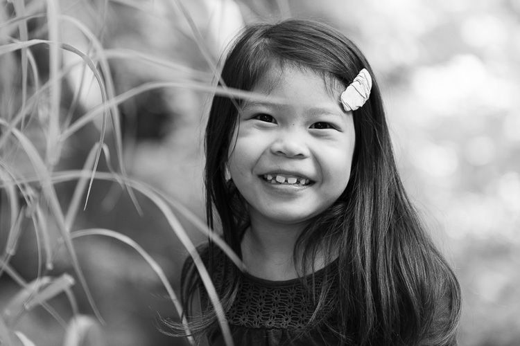 Black and white portrait of a smiling child with long dark hair and hair clips among tall grass, during a San Jose family photo session