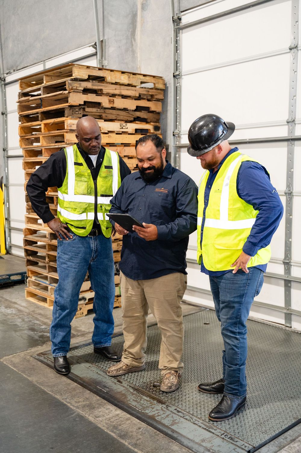 Workers in safety vests review documents on a tablet while standing near wooden pallets in a warehouse.