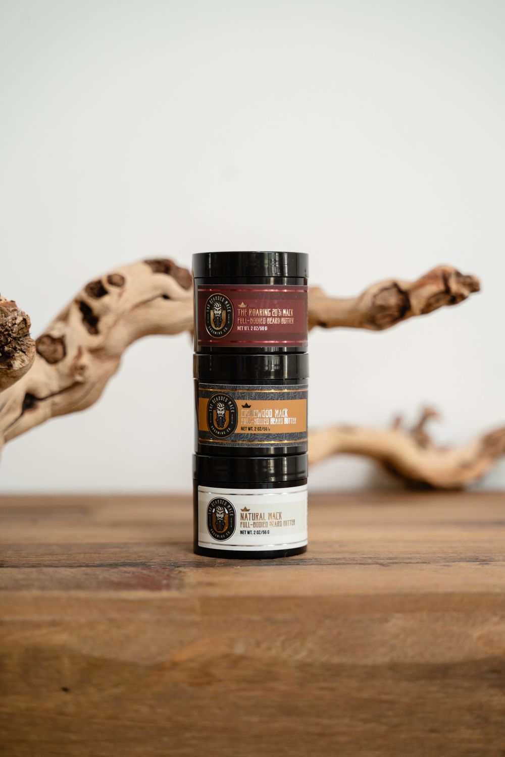 Small black cosmetic jars with gold labels arranged in a row on a rustic wooden surface with driftwood decor.