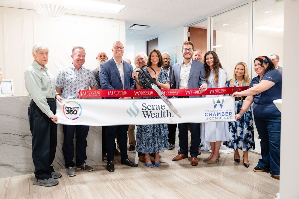 A group gathers for a ribbon cutting ceremony at a business opening with a large red ribbon spanning across the entrance.