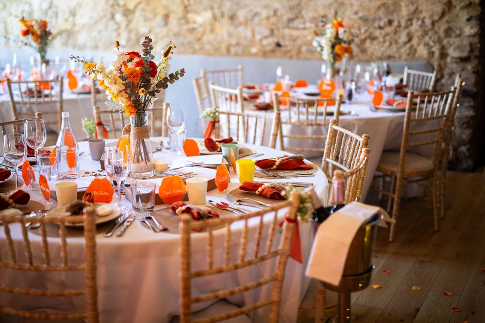 Elegant wedding reception tables decorated with orange accents and white chairs in rustic venue.