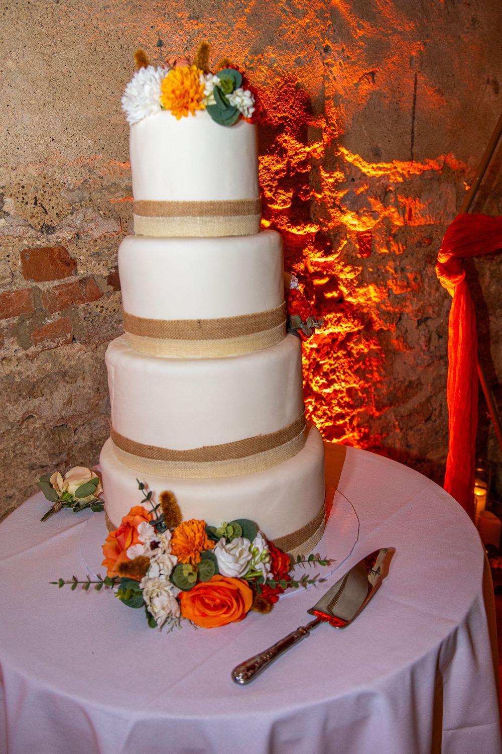 Four-tier white wedding cake with gold ribbon trim and orange floral decorations displayed against a dramatic fire-lit backdrop.