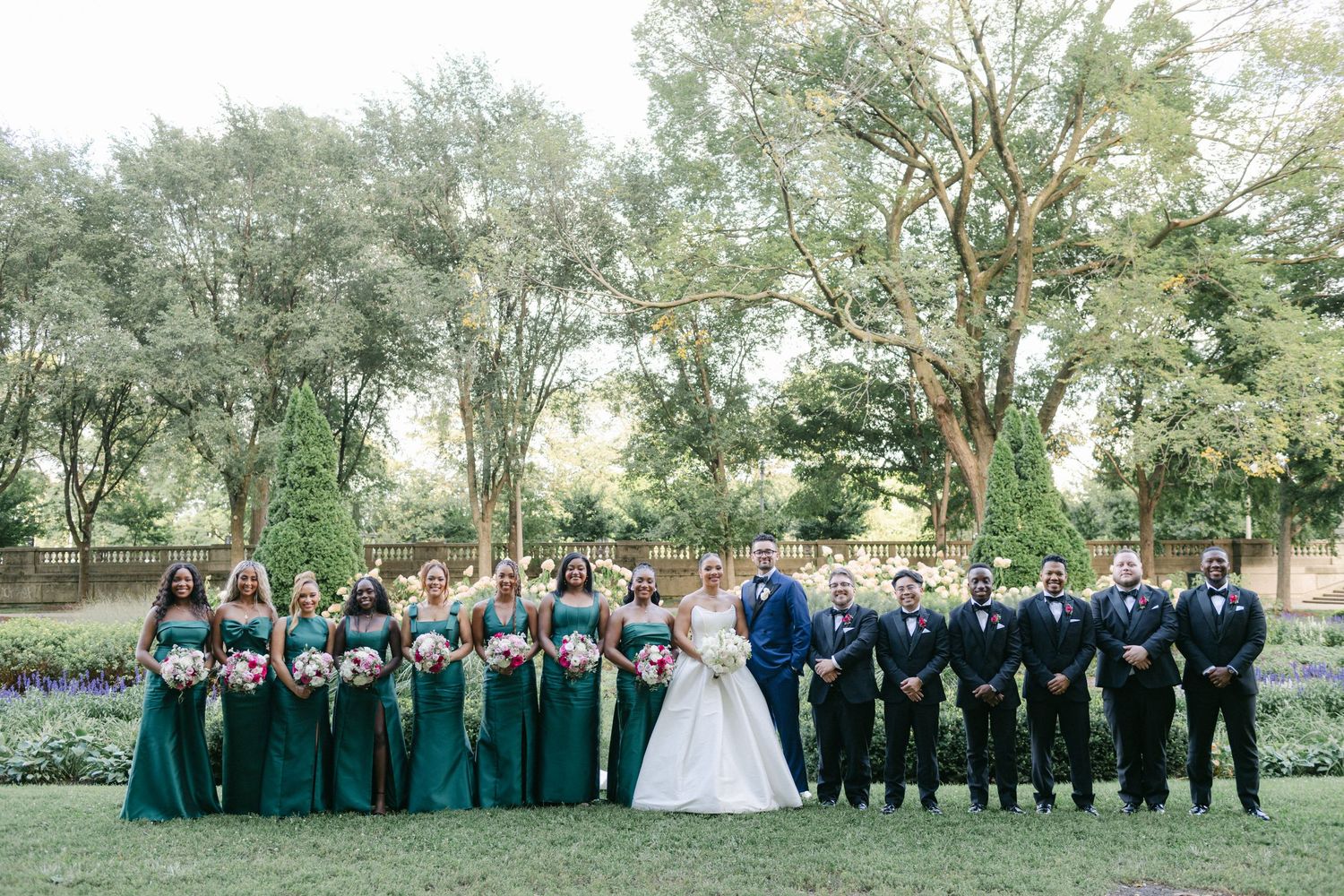 Wedding party poses outdoors in coordinating emerald green and black attire beneath tall trees at garden venue.