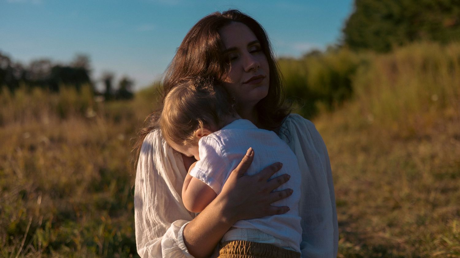 Child in white shirt explores wildflowers in a sunny meadow while someone sits in the background on a summer afternoon.