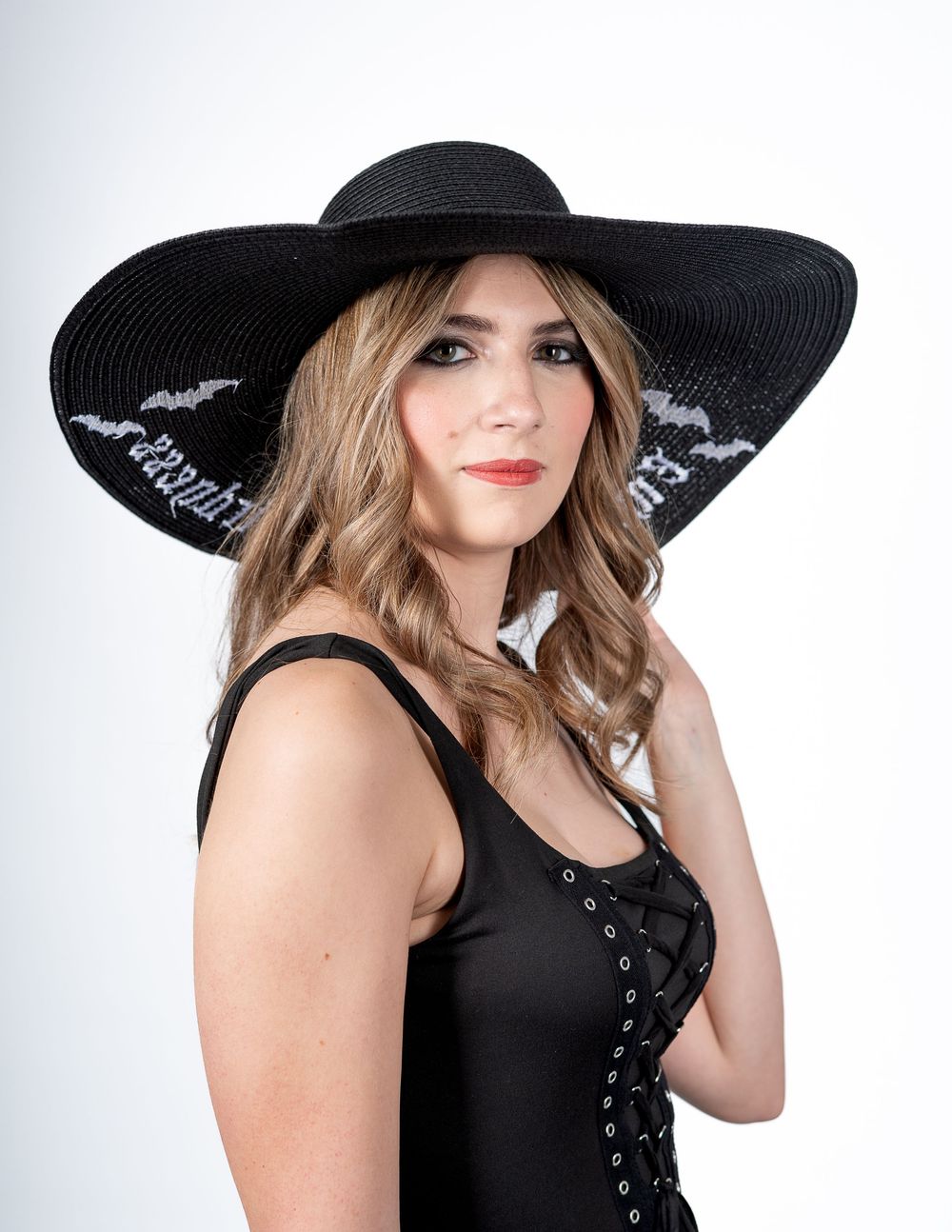 Model wearing a large black sunhat and black corset-style top poses elegantly against white background.
