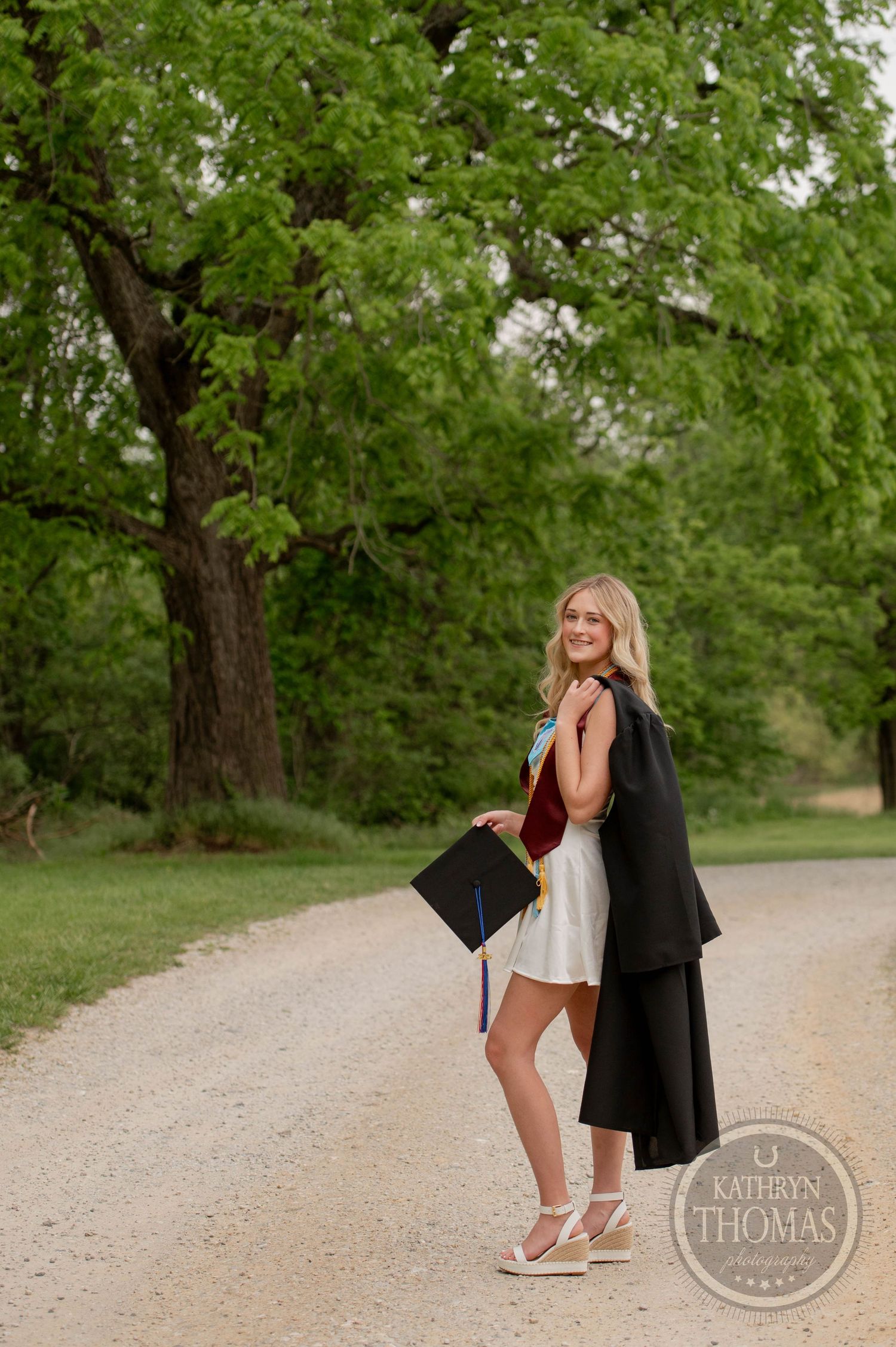 A graduate in a white dress holds up their graduation cap while standing on a dirt path under green trees.