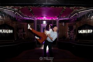 Two people dance intimately in a dark nightclub with purple lighting and mirrored walls.