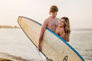A couple carrying surfboards walks along a beach at sunset during a romantic photoshoot.