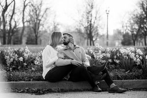 A couple sits together on a curb surrounded by blooming daffodils in a black and white romantic outdoor scene.