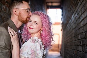 A couple embraces in a brick alley while the person with vibrant pink curly hair wears a lace wedding dress.