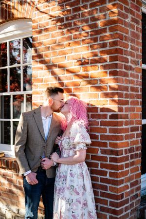 A couple in romantic pose against a sunlit brick wall, with soft shadows creating an intimate atmosphere.