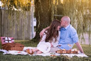 Two people share a romantic picnic on a blanket under a willow tree at sunset with woven baskets nearby.