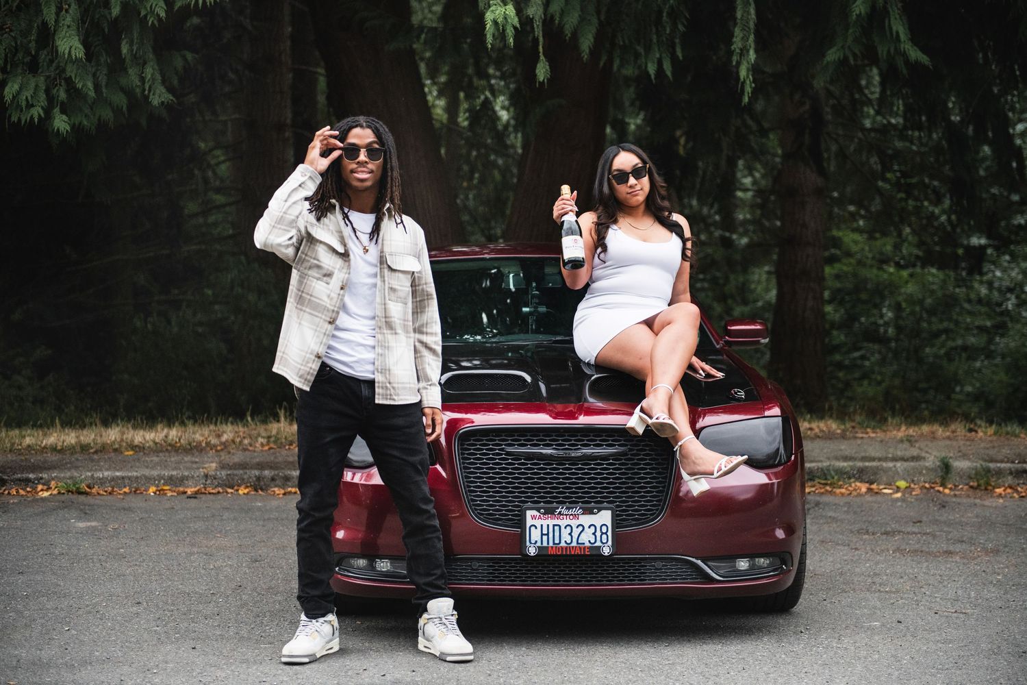 Two people pose with a burgundy car on a road surrounded by evergreen trees in a stylish lifestyle photo shoot.