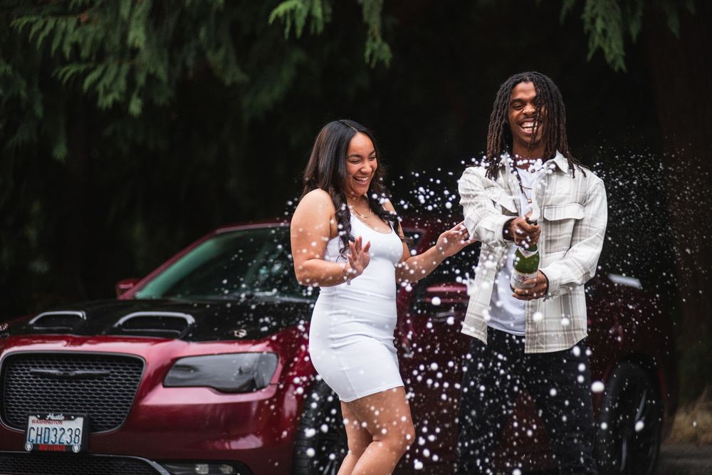 Two people celebrate joyfully spraying champagne while standing in front of a burgundy car.