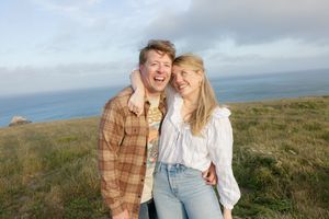 Couple sharing laughter against dramatic coastal landscape with ocean and cloudy sky.