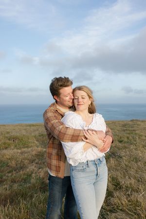 Intimate couple embraces on grassy coastal clifftop with ocean and cloudy sky in background.
