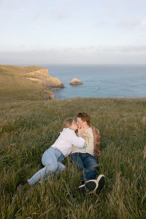 A couple shares an intimate moment while laying in tall grass on a coastal cliff overlooking the ocean at sunset.